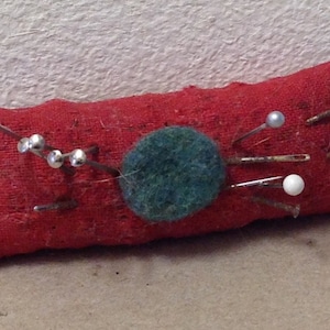 May include: A red fabric pincushion with a green felt circle in the center.  The pincushion is filled with pins with white and silver heads.