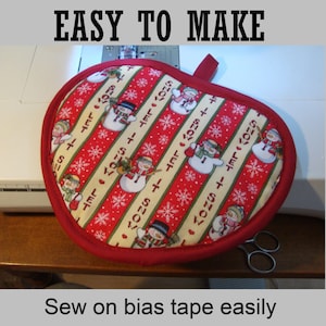 Puede incluir: Un agarrador de olla de tela rojo y blanco con un patrón de muñeco de nieve y el texto "Let it snow". El agarrador de olla está sobre una mesa de máquina de coser blanca con un par de tijeras plateadas.