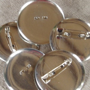 May include: Five blank, silver-coloured pinback buttons. Each button has a safety pin on the reverse for attachment. The buttons are arranged on a beige linen surface with a decorative lace trim.