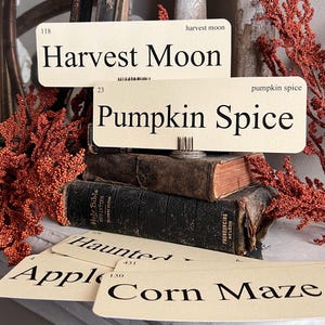 May include: A stack of vintage books with several cream-colored cards featuring fall-themed words like "Harvest Moon" and "Pumpkin Spice." The cards are arranged with a metal clip and surrounded by orange decorative branches.