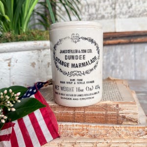 May include: A vintage James Keiller & Son Ltd. Dundee Orange Marmalade jar. The off-white ceramic jar has black text and a decorative border. The label reads "Net Weight 16oz (1lb) 454g". The jar sits on old books with a small American flag.