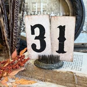 May include: Vintage-style display featuring two cards with the number "31" in black, set against a backdrop of aged books, feathers, and a vintage flower frog. The cards have a distressed, aged appearance.