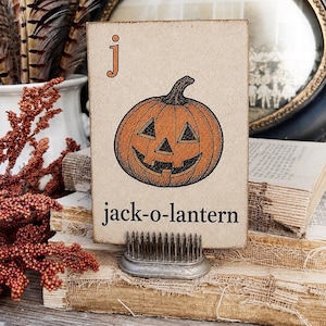 May include: A rectangular wooden sign featuring a printed illustration of an orange jack-o-lantern, the letter j in orange, and the black text jack-o-lantern. The sign is held upright by a metal pin frog stand with numerous vertical metal spikes.