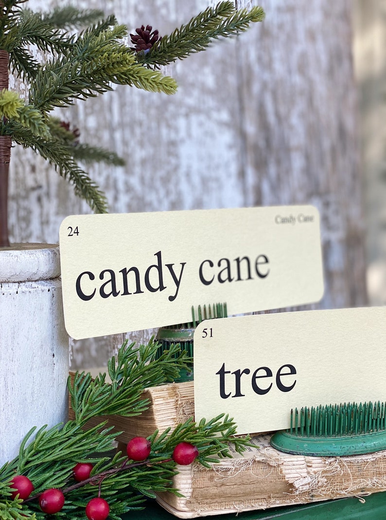 Peut inclure: Deux cartons cr&egrave;me avec le texte noir &laquo;&nbsp;candy cane&nbsp;&raquo; et &laquo;&nbsp;tree&nbsp;&raquo; sont affich&eacute;s. Les cartons sont soutenus par des crapaudines vertes. Une branche de verdure et des baies rouges sont au premier plan, avec un petit arbre de No&euml;l en arri&egrave;re-plan.