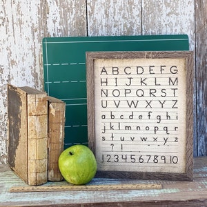 May include: A framed alphabet and number chart with a rustic wooden frame. Two antique books, a green apple, and a green chalkboard are next to the chart. A wooden ruler is at the bottom. The chart displays the letters A-Z and numbers 1-10.