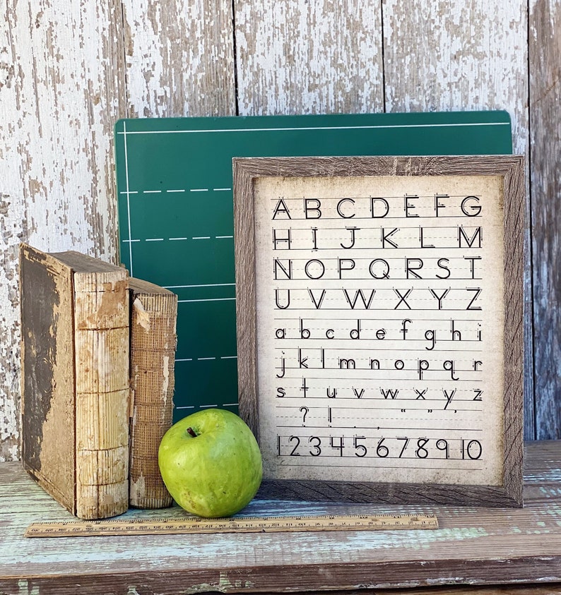 May include: A framed alphabet and number chart with a rustic wooden frame. Two antique books, a green apple, and a green chalkboard are next to the chart. A wooden ruler is at the bottom. The chart displays the letters A-Z and numbers 1-10.
