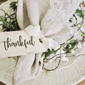 May include: A decorative place setting featuring a white plate with embossed floral patterns, a linen napkin, and a tag that reads "thankful". The tag is tied with a white ribbon and accented with greenery.