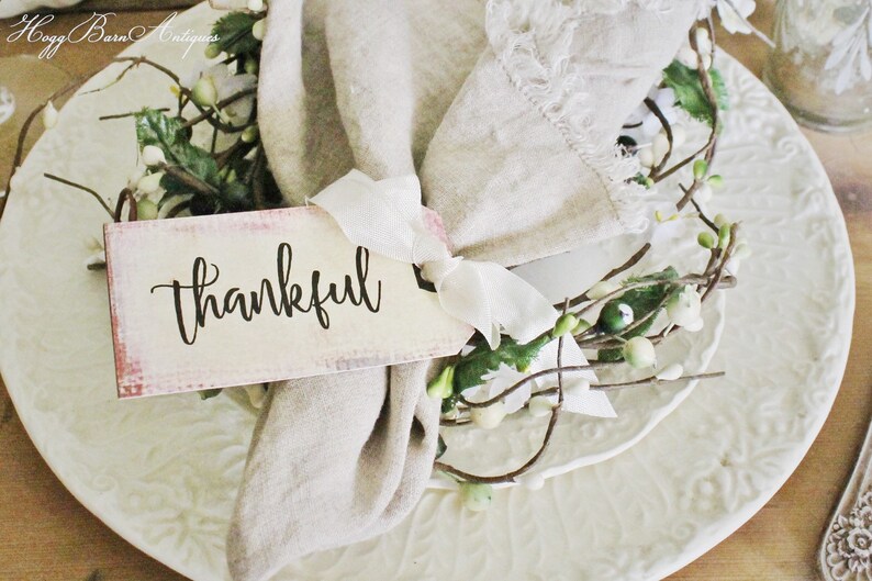 May include: A decorative place setting featuring a white plate with embossed floral patterns, a linen napkin, and a tag that reads "thankful". The tag is tied with a white ribbon and accented with greenery.