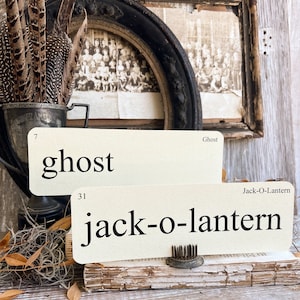 May include: Two cream-colored flashcards with the words "ghost" and "jack-o-lantern" printed in black. The cards are propped up on a stack of aged books. A vintage metal cup with feathers and a framed photograph are in the background.