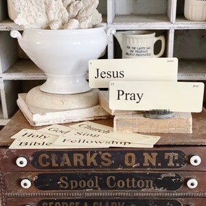 May include: A white ceramic urn filled with coral sits atop a stack of books, with index cards that read "Jesus" and "Pray". Below, a wooden drawer unit displays text that reads "CLARK'S O.N.T. Spool Cotton". The background features white cubby shelves.