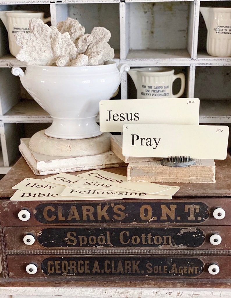 May include: A white ceramic urn filled with coral sits atop a stack of books, with index cards that read "Jesus" and "Pray". Below, a wooden drawer unit displays text that reads "CLARK'S O.N.T. Spool Cotton". The background features white cubby shelves.