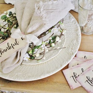 May include: A table setting with a white plate, a linen napkin, and a floral accent. A tag with the word "thankful" is tied to the napkin. Additional tags with the same word are on the table. A glass and silverware are also visible.