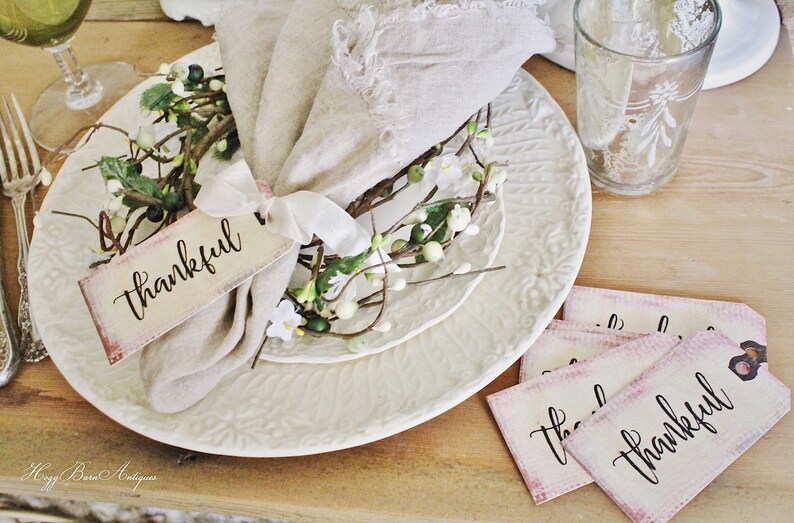 May include: A table setting with a white plate, a linen napkin, and a floral accent. A tag with the word "thankful" is tied to the napkin. Additional tags with the same word are on the table. A glass and silverware are also visible.