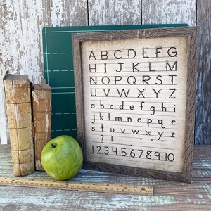 May include: A framed print with the alphabet and numbers, displayed with vintage books, a green apple, and a ruler. The print features uppercase and lowercase letters, along with numbers 1-10, in a handwritten style.