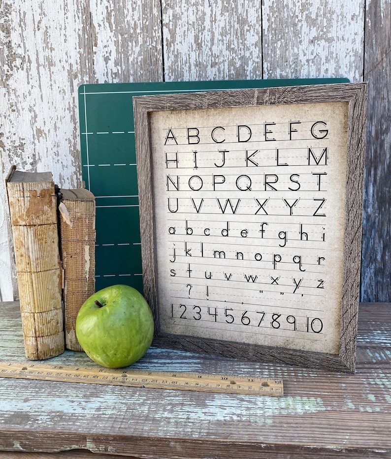 May include: A framed print with the alphabet and numbers, displayed with vintage books, a green apple, and a ruler. The print features uppercase and lowercase letters, along with numbers 1-10, in a handwritten style.