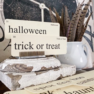 May include: A Halloween-themed display featuring vintage-style flashcards with words like "halloween" and "trick or treat." The cards are arranged with a white pitcher holding feathers, and a distressed wooden base. A large clock face is in the background.