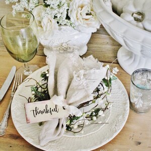 May include: A rustic table setting with a white plate, a linen napkin, and a tag that says "thankful." The plate is adorned with a small floral wreath. A green wine glass and a vase of white flowers complete the arrangement. The table is made of wood.