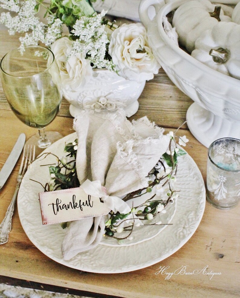 May include: A rustic table setting with a white plate, a linen napkin, and a tag that says "thankful." The plate is adorned with a small floral wreath. A green wine glass and a vase of white flowers complete the arrangement. The table is made of wood.