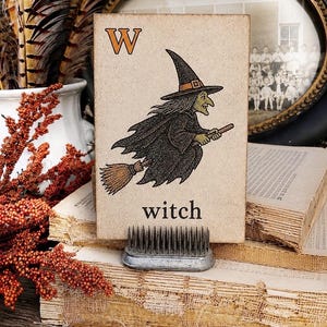 Puede incluir: Una tarjeta rectangular con la letra W en naranja y una ilustraci&oacute;n en tinta negra de una bruja volando sobre una escoba. La palabra witch est&aacute; impresa en letras min&uacute;sculas negras debajo de la ilustraci&oacute;n. La tarjeta se sostiene en un soporte.