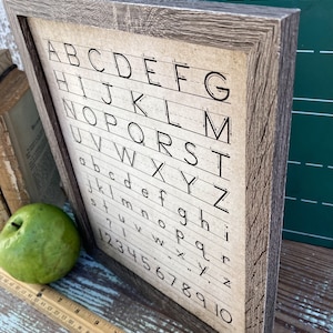 May include: Wooden framed sign featuring the alphabet and numbers 1-10 in a handwritten style. The sign has a rustic, weathered appearance with a brown frame and a beige background. A green apple and a ruler are next to the sign.