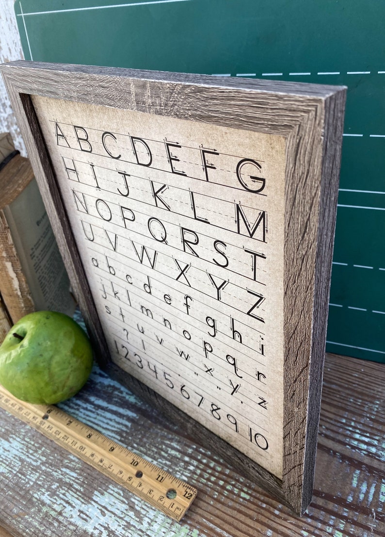 May include: Wooden framed sign featuring the alphabet and numbers 1-10 in a handwritten style. The sign has a rustic, weathered appearance with a brown frame and a beige background. A green apple and a ruler are next to the sign.