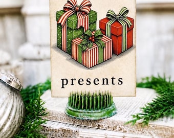 Puede incluir: Una tarjeta rectangular con la letra p y la palabra presents en texto negro, con una ilustración de tres cajas de regalo rojas y verdes con lazos. La tarjeta se mantiene en posición vertical mediante una base redonda de metal verde.