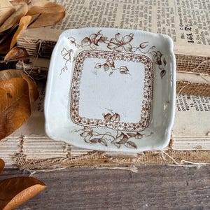 May include: A square ceramic dish with rounded corners, featuring a cream-colored base and a brown floral transferware pattern. The design includes a decorative border of intricate scrollwork and small flowers along the edges.