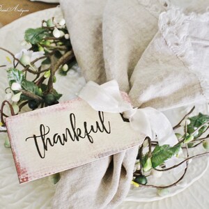 May include: A decorative place setting featuring a white plate with embossed detailing, a beige linen napkin, and a tag that reads "thankful." The tag is tied with a white ribbon and adorned with a small floral arrangement.