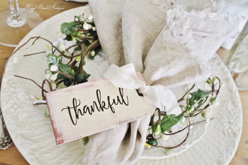May include: A decorative place setting featuring a white plate with embossed detailing, a beige linen napkin, and a tag that reads "thankful." The tag is tied with a white ribbon and adorned with a small floral arrangement.