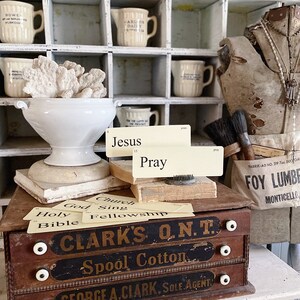 May include: A vintage still life with a white ceramic bowl holding coral, books, and cards with the words "Jesus" and "Pray". A wooden box with the text "CLARK'S O.N.T. Spool Cotton" is in the foreground. Various cream-colored jugs are in the background.