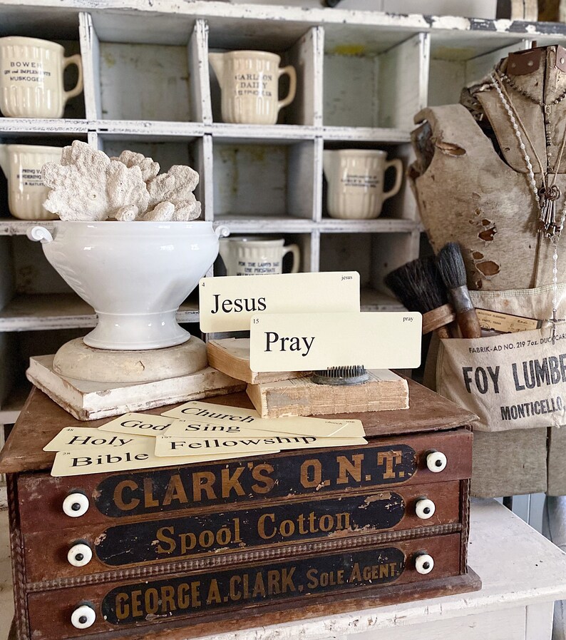 May include: A vintage still life with a white ceramic bowl holding coral, books, and cards with the words "Jesus" and "Pray". A wooden box with the text "CLARK'S O.N.T. Spool Cotton" is in the foreground. Various cream-colored jugs are in the background.