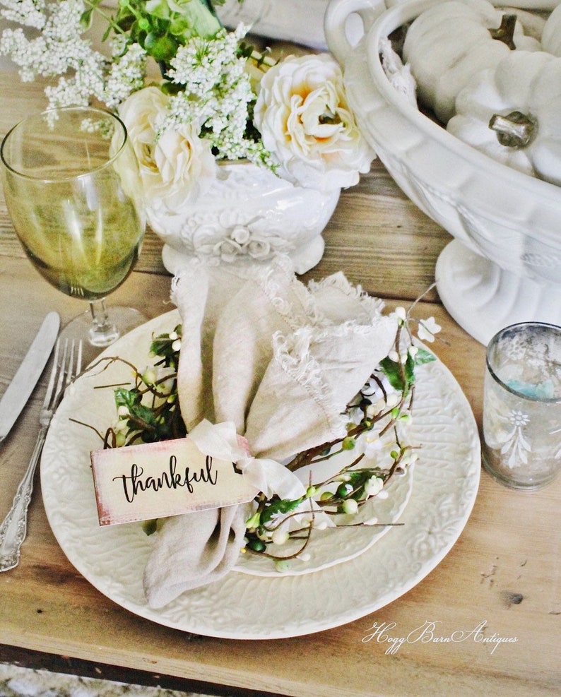 May include: A rustic table setting with a white plate, linen napkin, and floral accents. A tag with the word "thankful" is tied to the napkin. A wine glass and a vase of flowers are also present, creating a warm, inviting atmosphere.