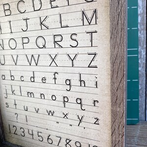 May include: A framed vintage-style alphabet and number chart. The chart features uppercase and lowercase letters, along with numbers 1-10, all in black ink on a beige background. The wooden frame has a rustic, weathered appearance.