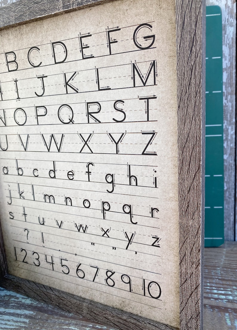 May include: A framed vintage-style alphabet and number chart. The chart features uppercase and lowercase letters, along with numbers 1-10, all in black ink on a beige background. The wooden frame has a rustic, weathered appearance.