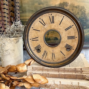 May include: A circular clock face with a dark metal rim and a weathered, beige background featuring black Roman numerals. The center has a circular cutout, and two small circular openings are positioned near the bottom, with a small diamond-shaped logo.