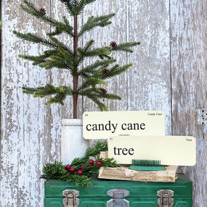 Peut inclure: Un petit sapin de No&euml;l artificiel avec des pommes de pin dans un pot blanc, pos&eacute; sur une bo&icirc;te &agrave; outils en m&eacute;tal vert. Deux cartons vintage indiquent "candy cane" et "tree". Une branche de verdure et des baies rouges sont &eacute;galement pr&eacute;sentes.