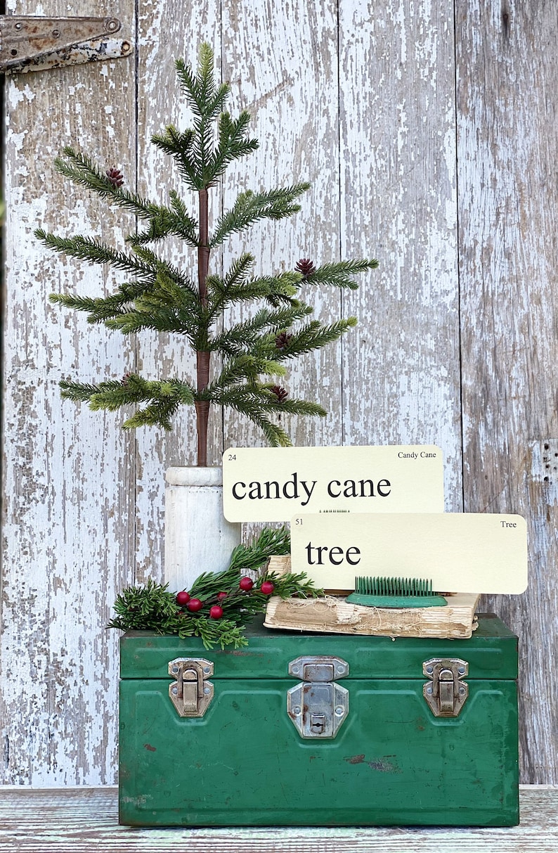 Peut inclure: Un petit sapin de No&euml;l artificiel avec des pommes de pin dans un pot blanc, pos&eacute; sur une bo&icirc;te &agrave; outils en m&eacute;tal vert. Deux cartons vintage indiquent "candy cane" et "tree". Une branche de verdure et des baies rouges sont &eacute;galement pr&eacute;sentes.