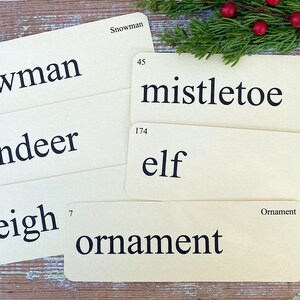 Peut inclure: Ensemble de cartes de No&euml;l vintage avec les mots : bonhomme de neige, gui, renne, elfe, tra&icirc;neau et ornement. Les cartes cr&egrave;me avec texte noir sont dispos&eacute;es sur une surface en bois rustique, avec des branches de conif&egrave;res et des baies rouges.