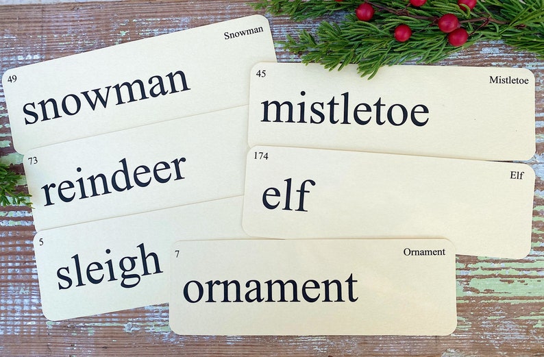 Peut inclure: Ensemble de cartes de No&euml;l vintage avec les mots : bonhomme de neige, gui, renne, elfe, tra&icirc;neau et ornement. Les cartes cr&egrave;me avec texte noir sont dispos&eacute;es sur une surface en bois rustique, avec des branches de conif&egrave;res et des baies rouges.