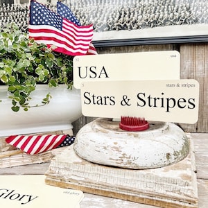 May include: A patriotic display featuring an American flag, a vintage-style sign that reads "USA Stars & Stripes", and a red flower frog. The items are arranged on a distressed wooden base, evoking a rustic aesthetic.