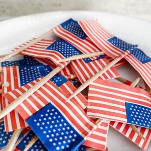 May include: A collection of small American flags with red and white stripes and blue fields of stars. The flags are attached to wooden sticks and are arranged on a white surface, likely for a patriotic celebration or decoration.
