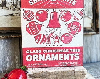 May include: A vintage red and white "Shiny Brite" Christmas ornament box. The box features illustrations of ornaments and the text "Glass Christmas Tree Ornaments." A red ornament and greenery are in the foreground.