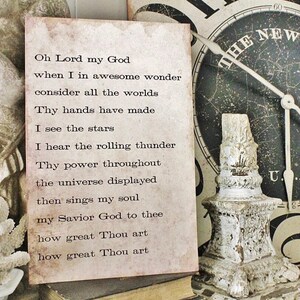 May include: A vintage-style print with the text of a hymn, "Oh Lord my God." The print has a distressed, aged appearance with brown and beige tones. A decorative, white, ornate candle holder and a clock with the text "The New Era" are in the background.