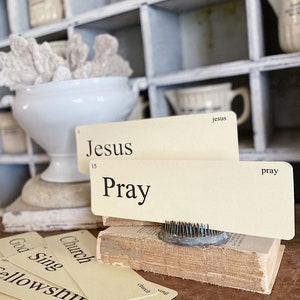 May include: A still life composition with vintage flashcards bearing religious terms such as "Jesus" and "Pray." The cards are arranged on a small, aged book, with a metal flower frog. A white ceramic bowl with coral is in the background.