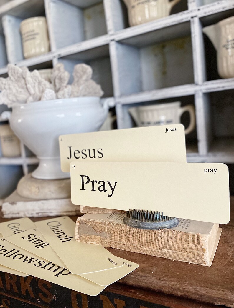 May include: A still life composition with vintage flashcards bearing religious terms such as "Jesus" and "Pray." The cards are arranged on a small, aged book, with a metal flower frog. A white ceramic bowl with coral is in the background.