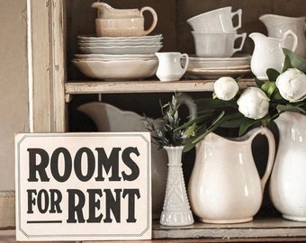 Peut inclure: Un buffet en bois rustique présente de la vaisselle en céramique blanche, des pichets et des tasses. Une enseigne indiquant « ROOMS FOR RENT » est au premier plan. L'ensemble évoque une esthétique vintage, avec des plats de différentes tailles.