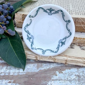 May include: A small, white ceramic dish with a decorative teal border. The border features an ornate, repeating pattern of stylized floral and scroll designs. The dish is set on a stack of aged paper, suggesting a vintage or antique aesthetic.