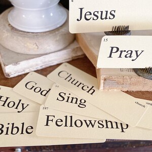 May include: Cream flash cards with black text, featuring words such as "Jesus," "Pray," and "Bible." The cards are displayed on a wooden surface with a vintage look. A white ceramic object and a book are in the background.