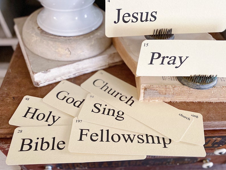 May include: Cream flash cards with black text, featuring words such as "Jesus," "Pray," and "Bible." The cards are displayed on a wooden surface with a vintage look. A white ceramic object and a book are in the background.
