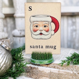 Op de afbeelding: Een rechthoekige beige kaart met de letter S bovenaan en een gedetailleerde illustratie van een mok in de vorm van een kerstmangezicht met een rood handvat in het midden. De tekst santa mug staat in het zwart onderaan de rechtopstaande kaart.
